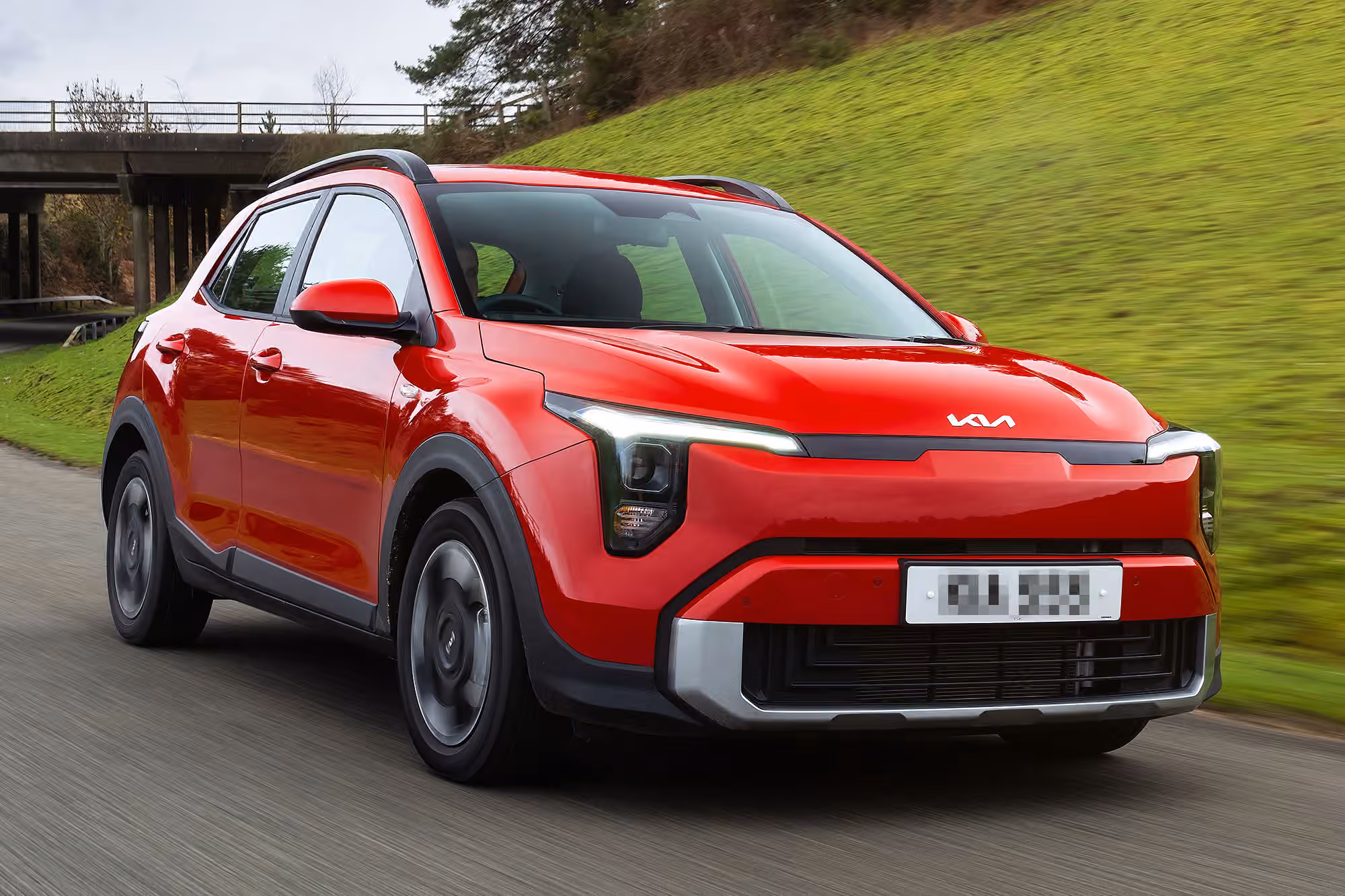 Red Kia crossover driving on a country road with green hillside and bridge in the background.