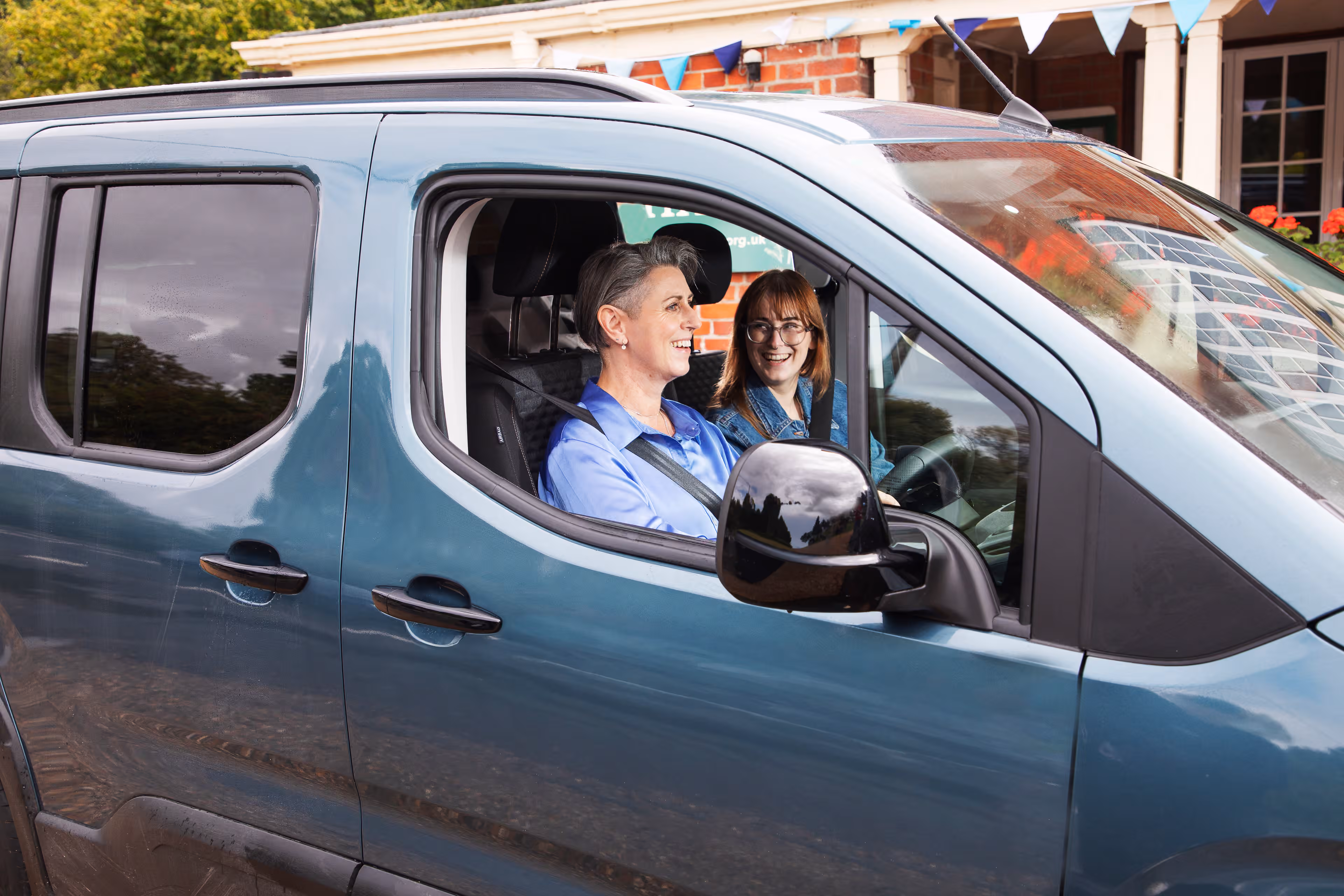 Two smiling women in blue shirts sitting in a teal van parked outside a brick house with bunting decorations.