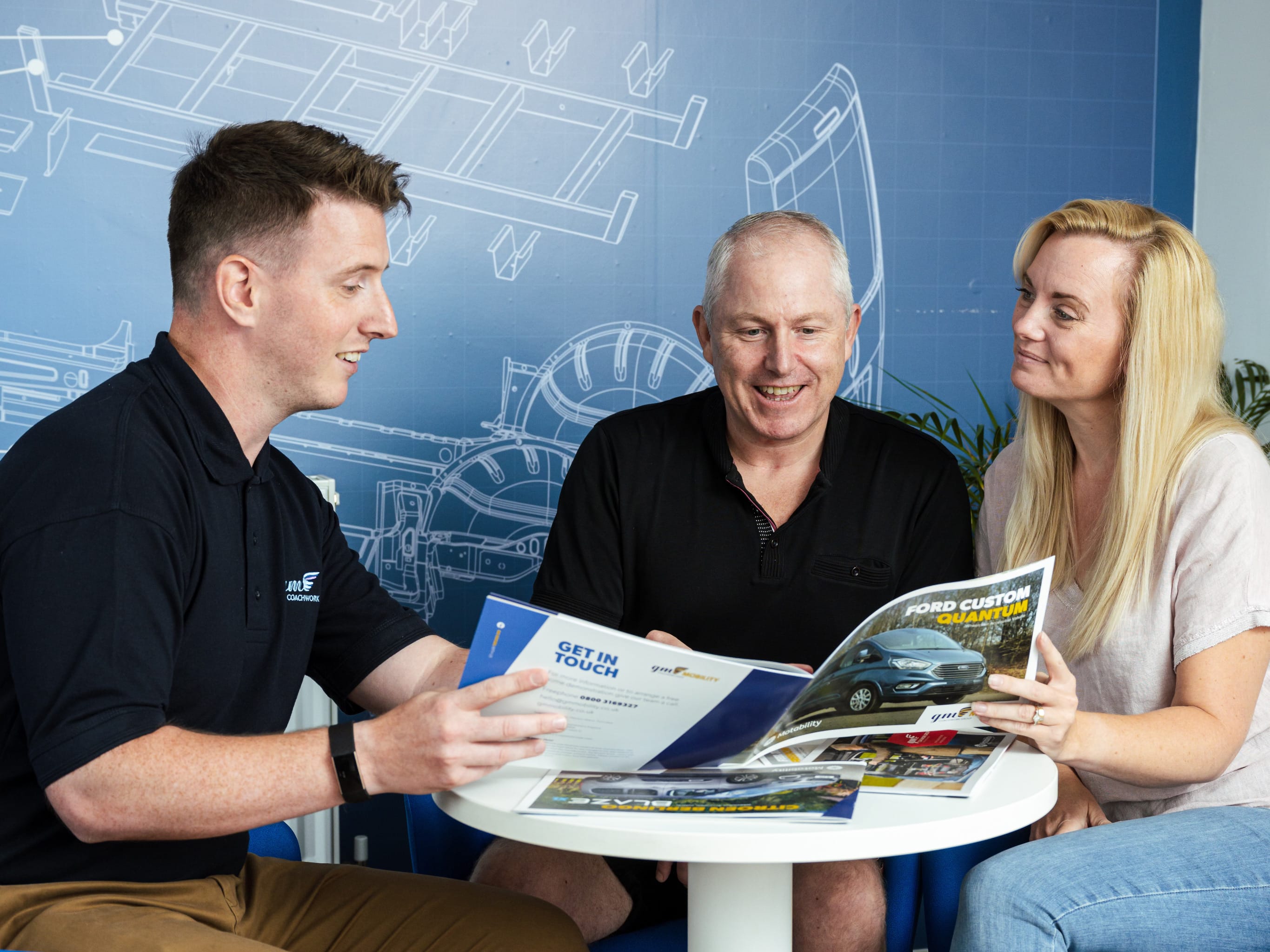 A vehicle dealer and two people reviewing Ford brochures at a white table against a blue wall with vehicle blueprint illustrations.