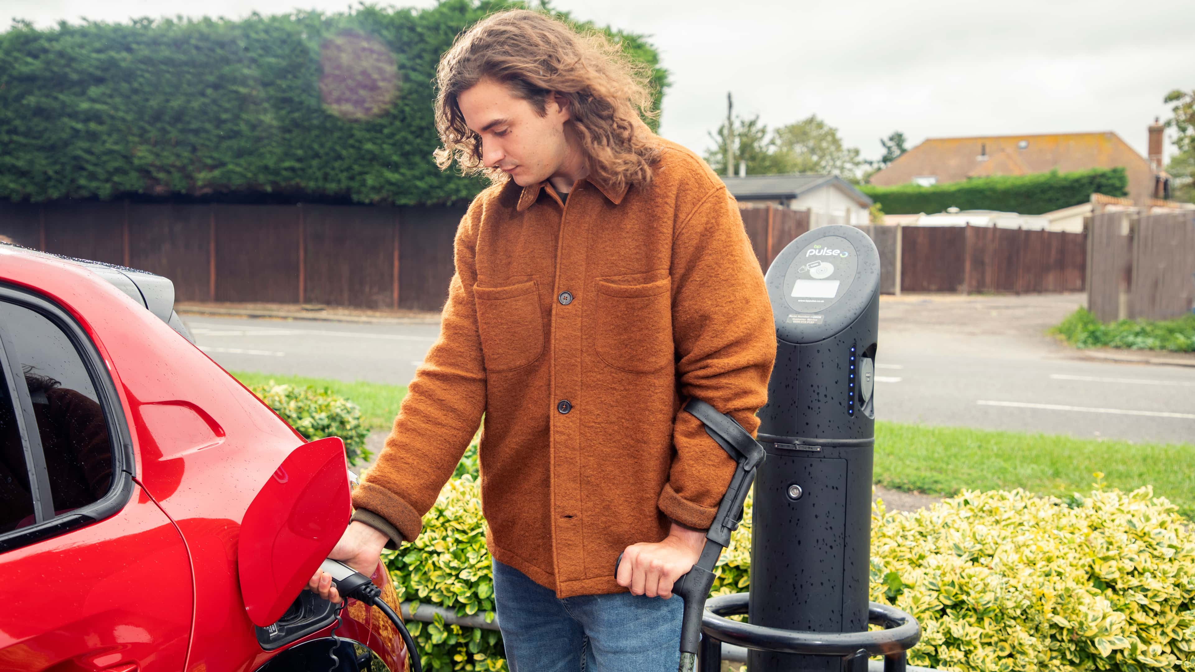 A man in a rust-coloured jacket charges a red electric car at a roadside EV charging point, holding a walking aid under one arm.