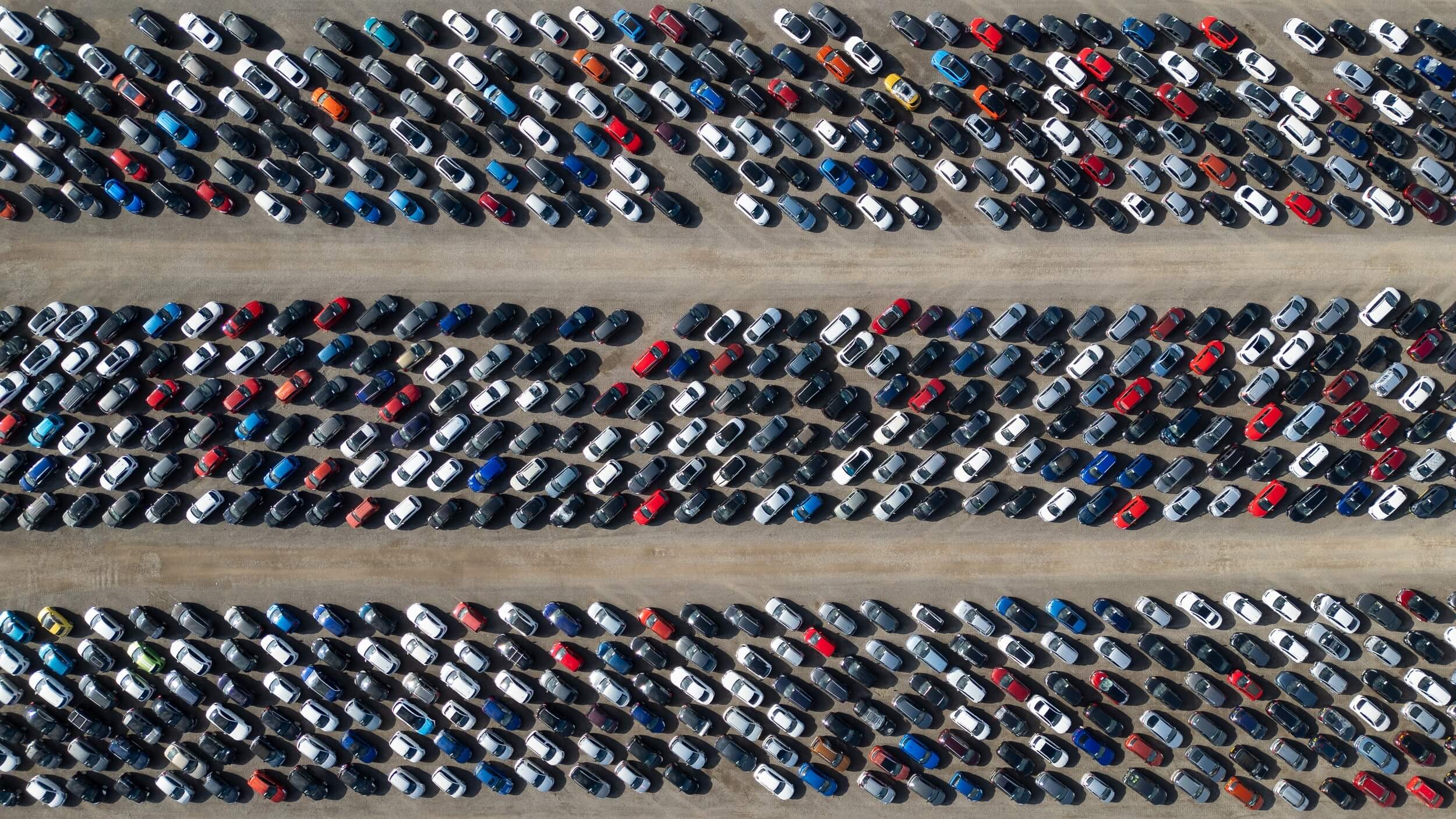 Aerial view of a large outdoor car park with hundreds of cars parked in long, diagonal rows separated by wide access lanes. Most vehicles are white, black, grey or blue, with a few red, orange and yellow cars adding contrast. The image fills the frame with repeating patterns of tightly parked vehicles, viewed from directly above.